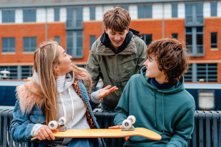 Two teen boys with one teen girl smiling and talking while two of them hold a skateboard