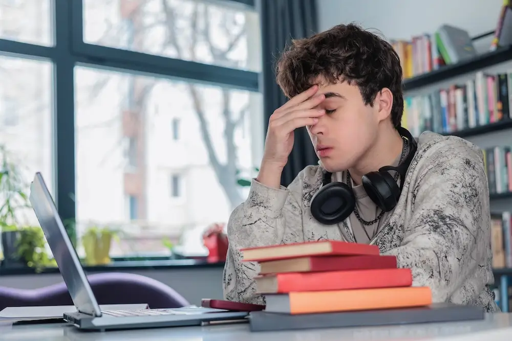 Teenage boy sitting at desk with computer and school books with eyes closed and hand on forehead
